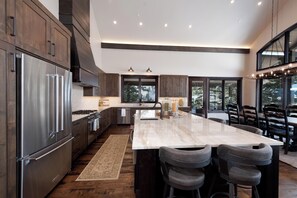 A modern kitchen with dark wood cabinets, stainless steel appliances, a large island with bar stools, and large windows offering a view of trees.