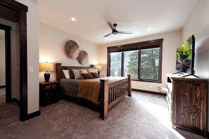 A bedroom with a large bed, a ceiling fan, a window with a view of trees, and a wooden cabinet.