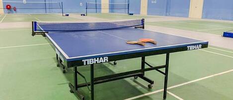 Ett bordtennisbord för inomhusbruk med en blå spelyta och ett nät, märkt med texten ”TIBHAR”, i en sportanläggning med grönt golv och blå väggar.