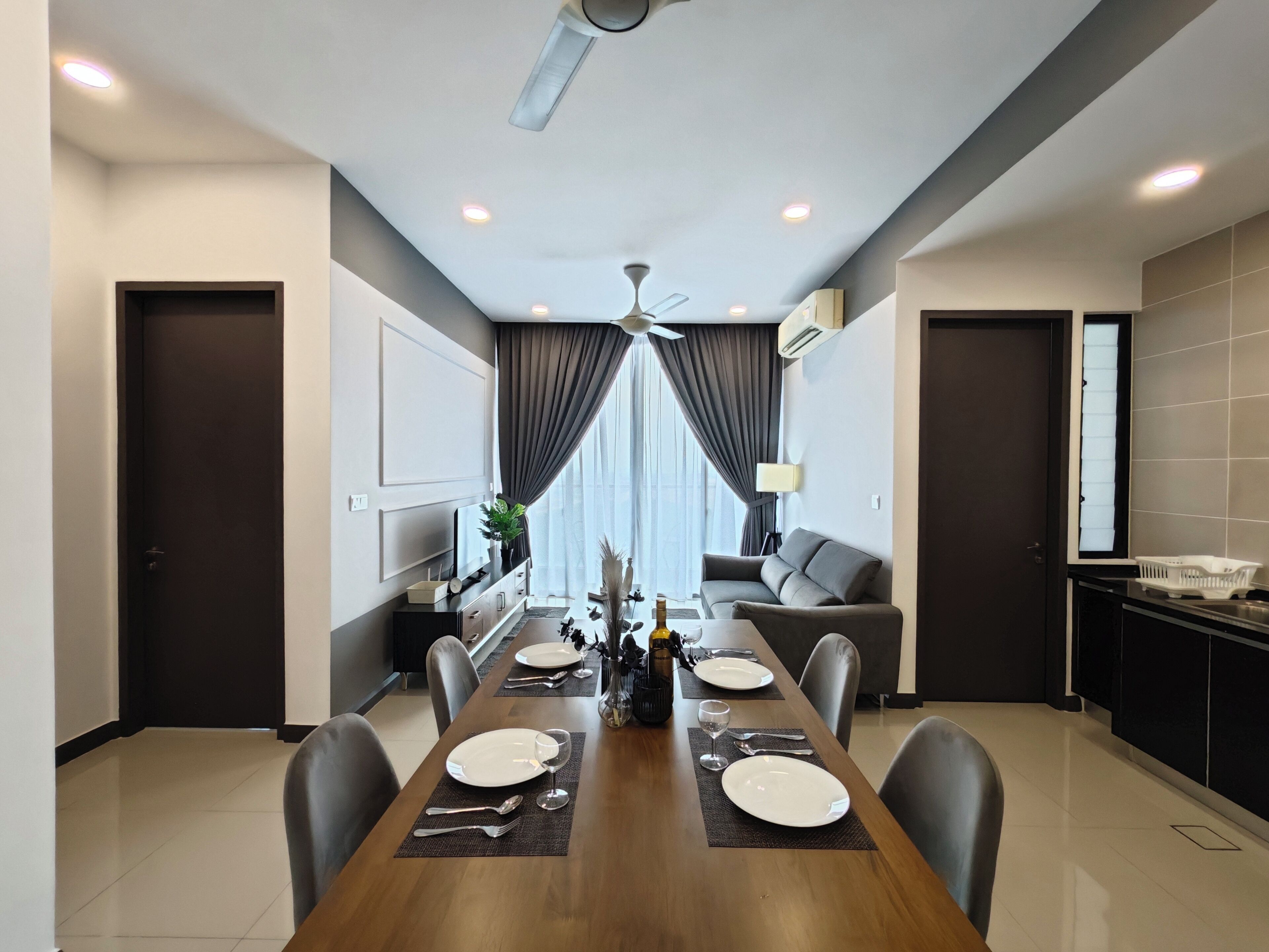 A modern dining area with a wooden table set for four, flanked by dark sofas and a kitchen area.