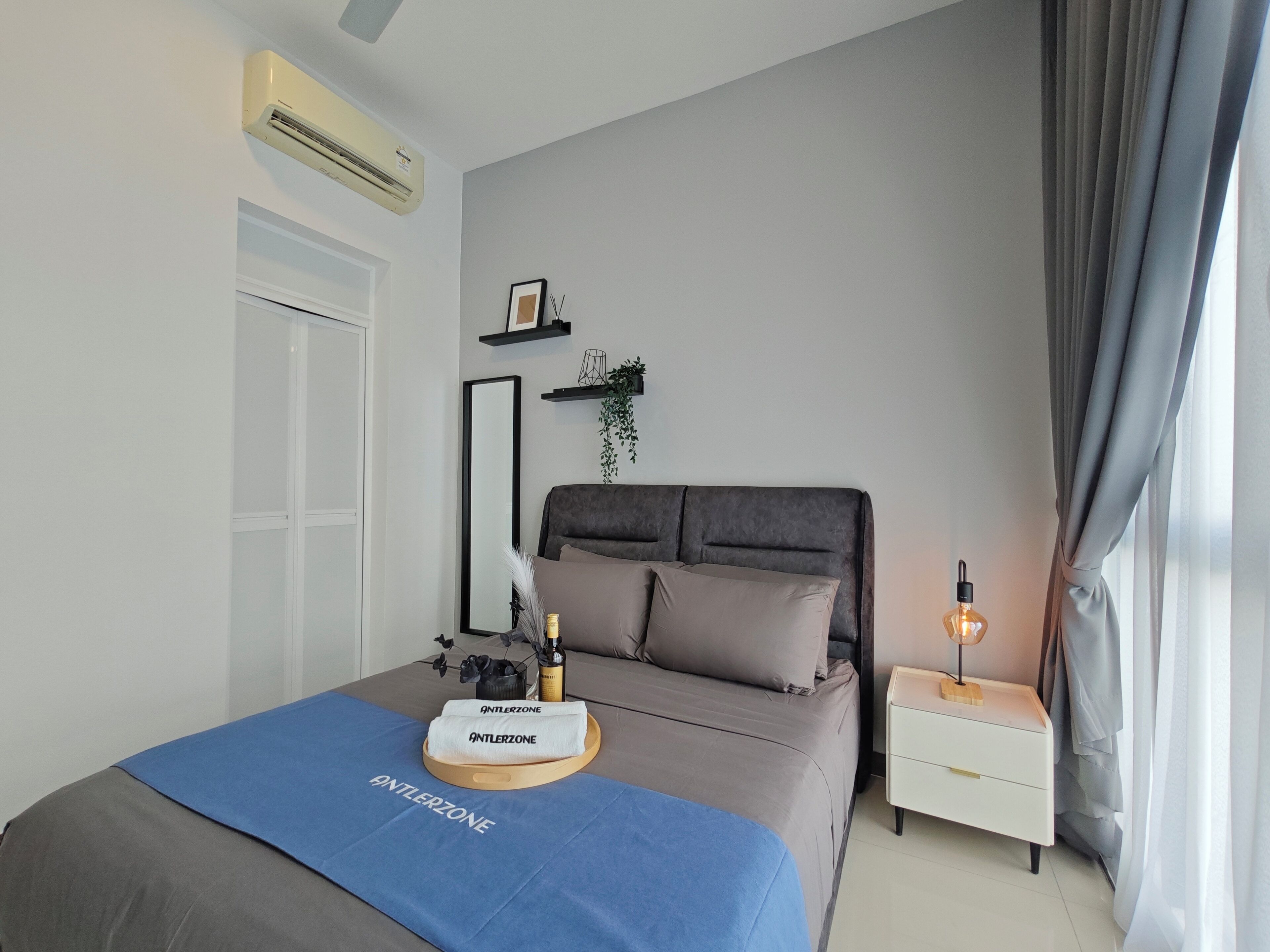 A modern bedroom with a grey bed, a bedside table with a lamp, a blue towel, and a small plant.