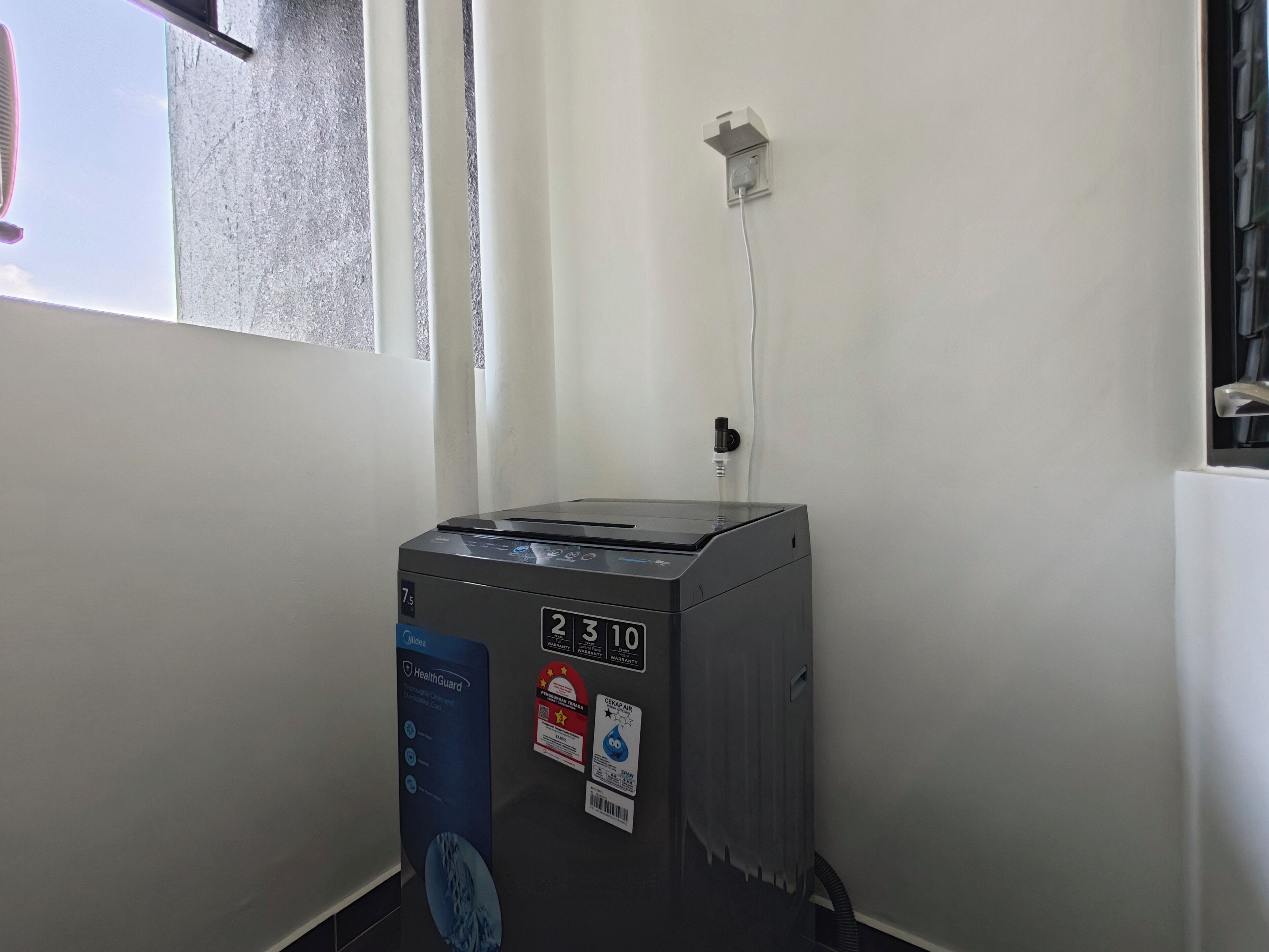 Un purificador de aire compacto y moderno con pantalla digital, ubicado en una esquina de una habitación con pared blanca lisa y una ventana.