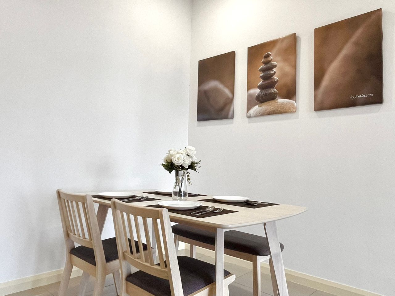 Un comedor con una mesa de madera, cuatro sillas, un florero con flores blancas y tres cuadros en la pared.