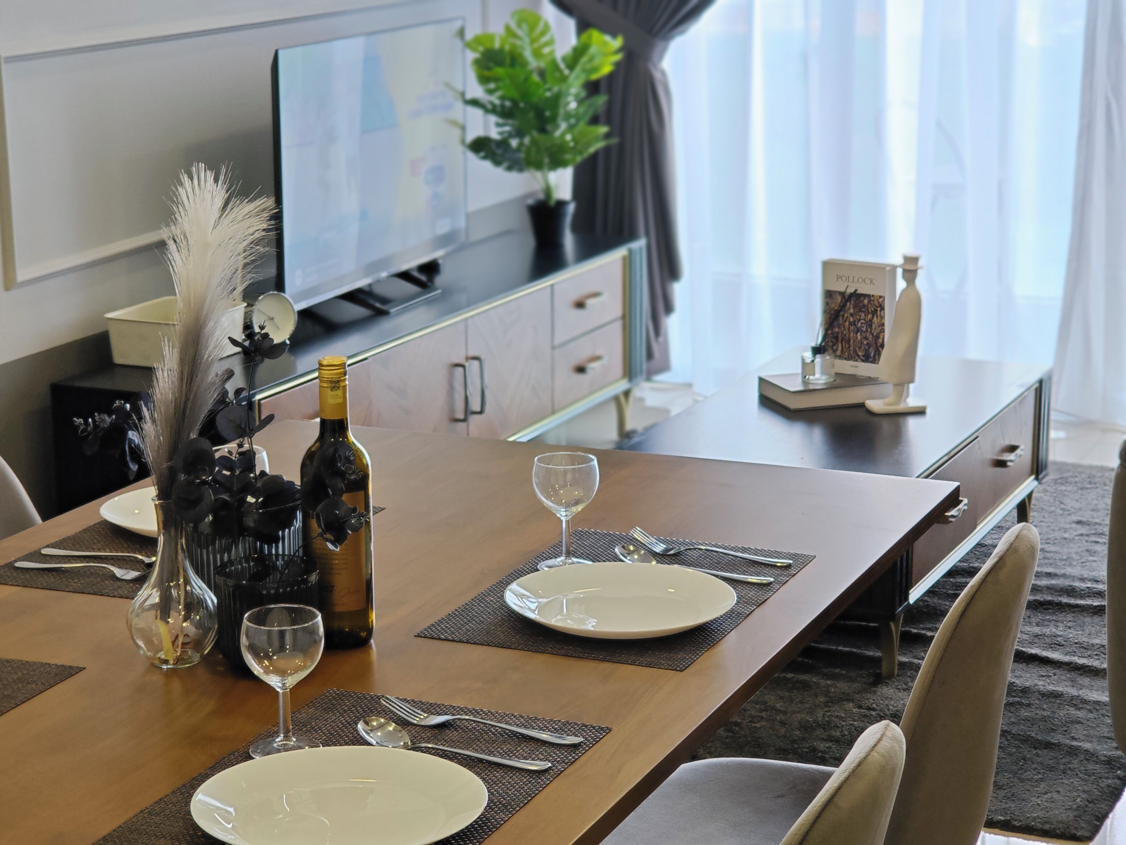 Una mesa de comedor dispuesta con platos, cubiertos y una botella de vino, con un televisor y una planta al fondo.