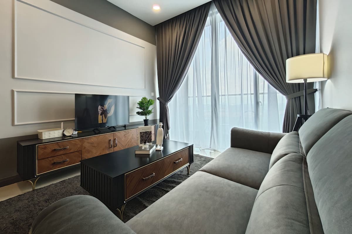 Ein modernes Wohnzimmer mit einem Flachbildfernseher, einer dunklen Holzkonsole, einem grauen Sofa und einem großen Fenster mit Vorhängen.
