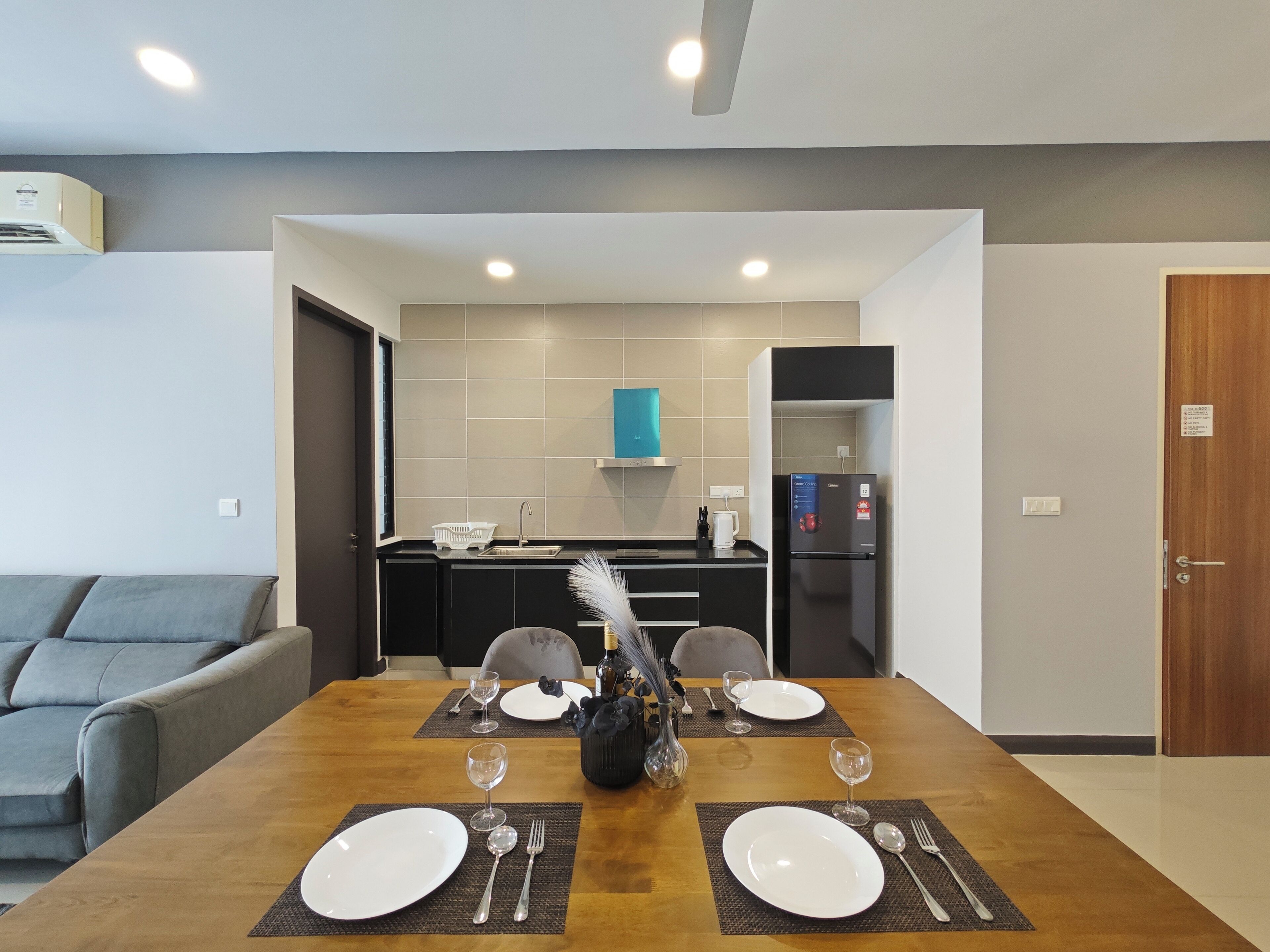 A modern dining room with a wooden table set for four, a kitchen in the background, and a grey sofa.