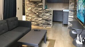 Ein modernes Wohnzimmer mit einer Couch, einem Couchtisch und einem Fernseher. Es gibt eine Küchenzeile mit Schränken und einer Mikrowelle.