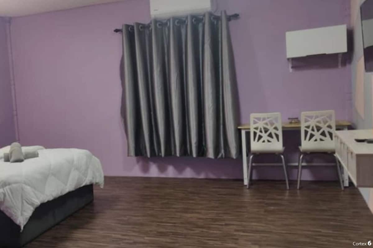 A room with a bed, a desk, two chairs, a window with curtains, and a wall-mounted fan.