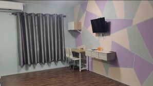 A room with a wooden floor, a desk with a chair, a television mounted on the wall, and a window with curtains.