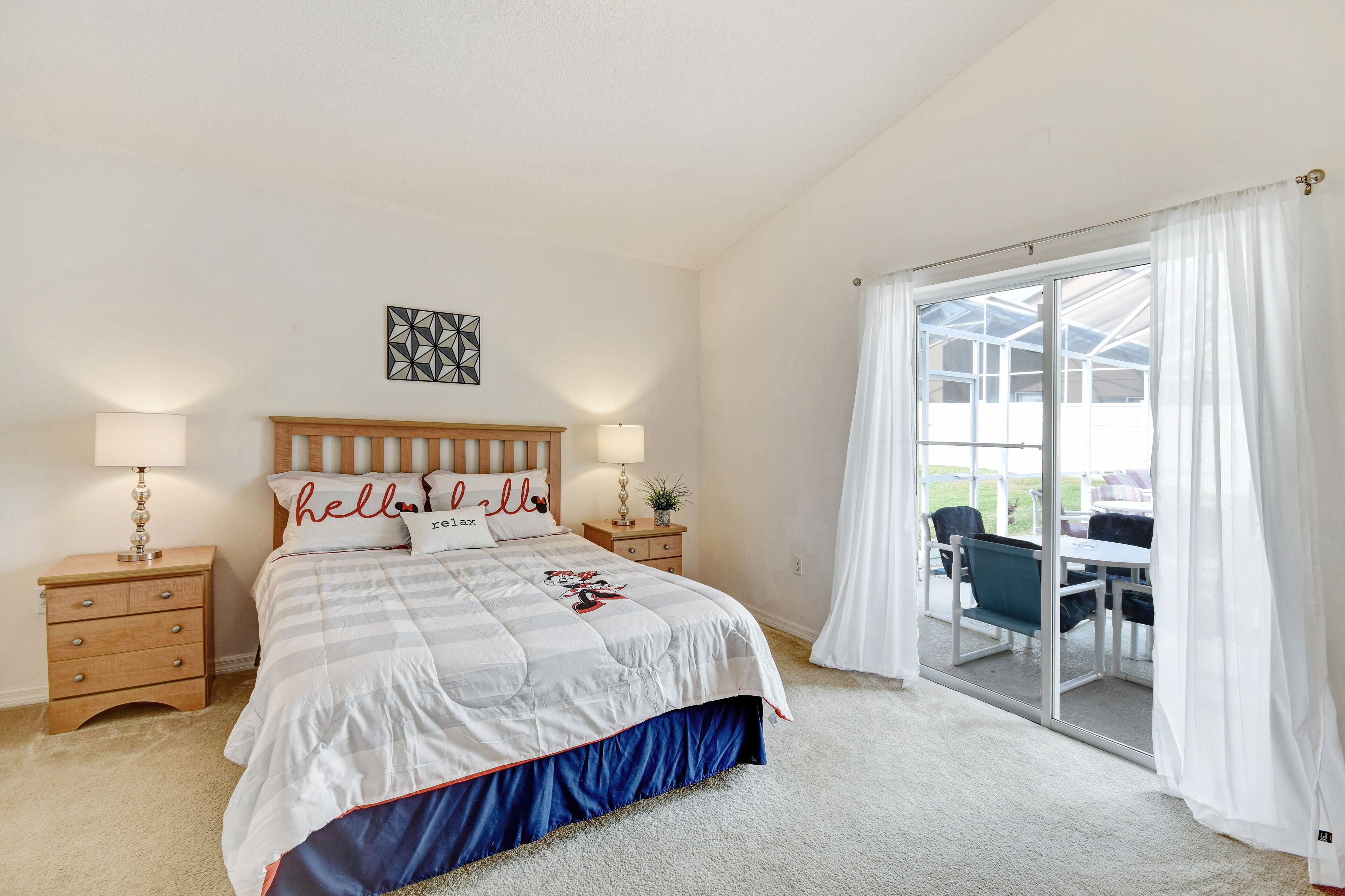 A bedroom with a large bed, two bedside tables with lamps, a headboard with decorative pillows, and a view through a sliding glass door to an outdoor area.