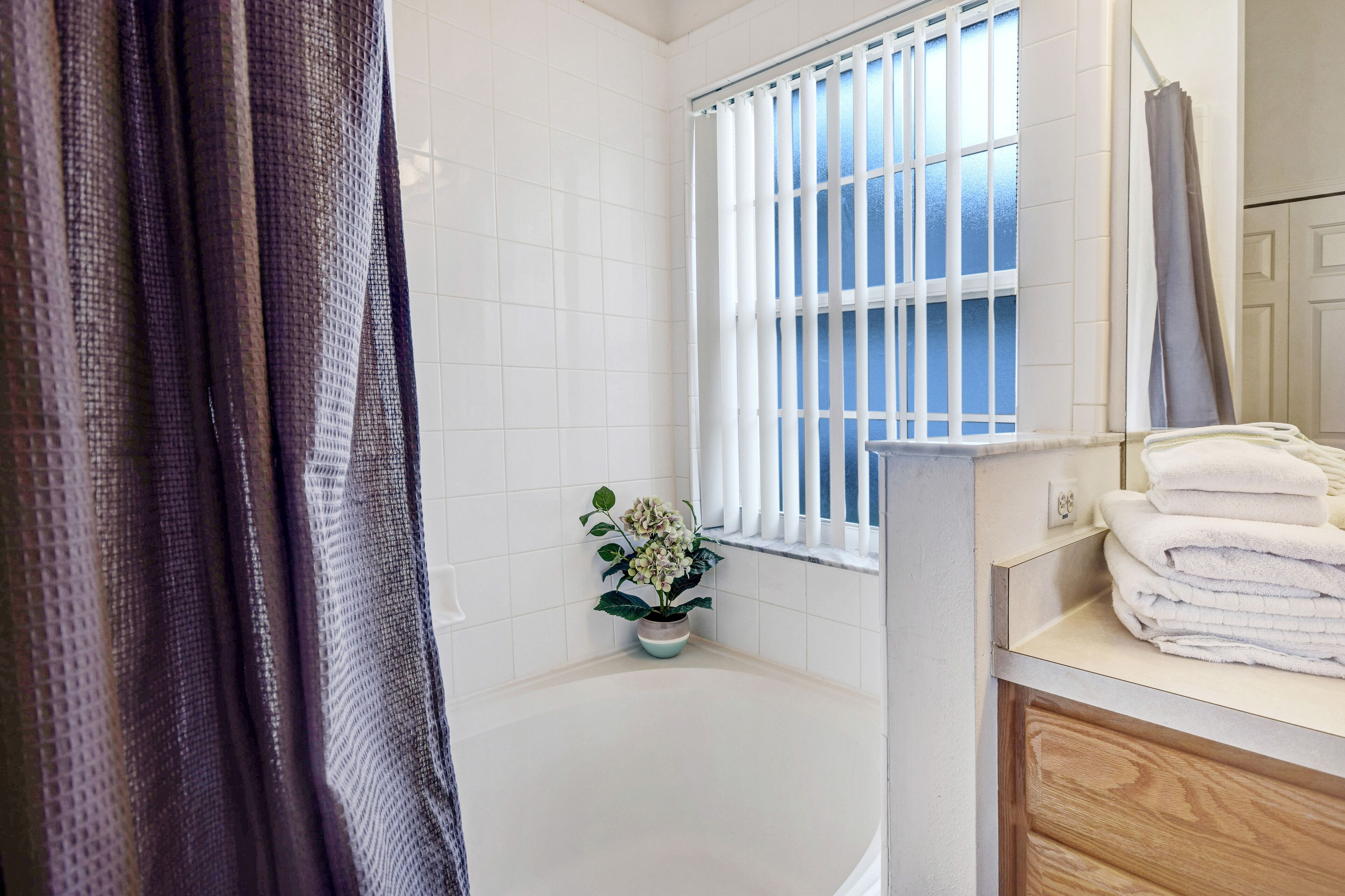 A bathroom with a bathtub, a window with blinds, a potted plant, and a stack of towels.