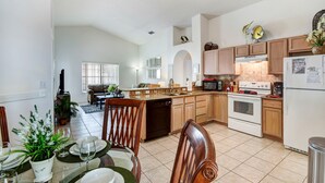 A spacious kitchen with wooden cabinets, a dishwasher, a stove, and a refrigerator. There is a dining area with a table set for two and a potted plant.