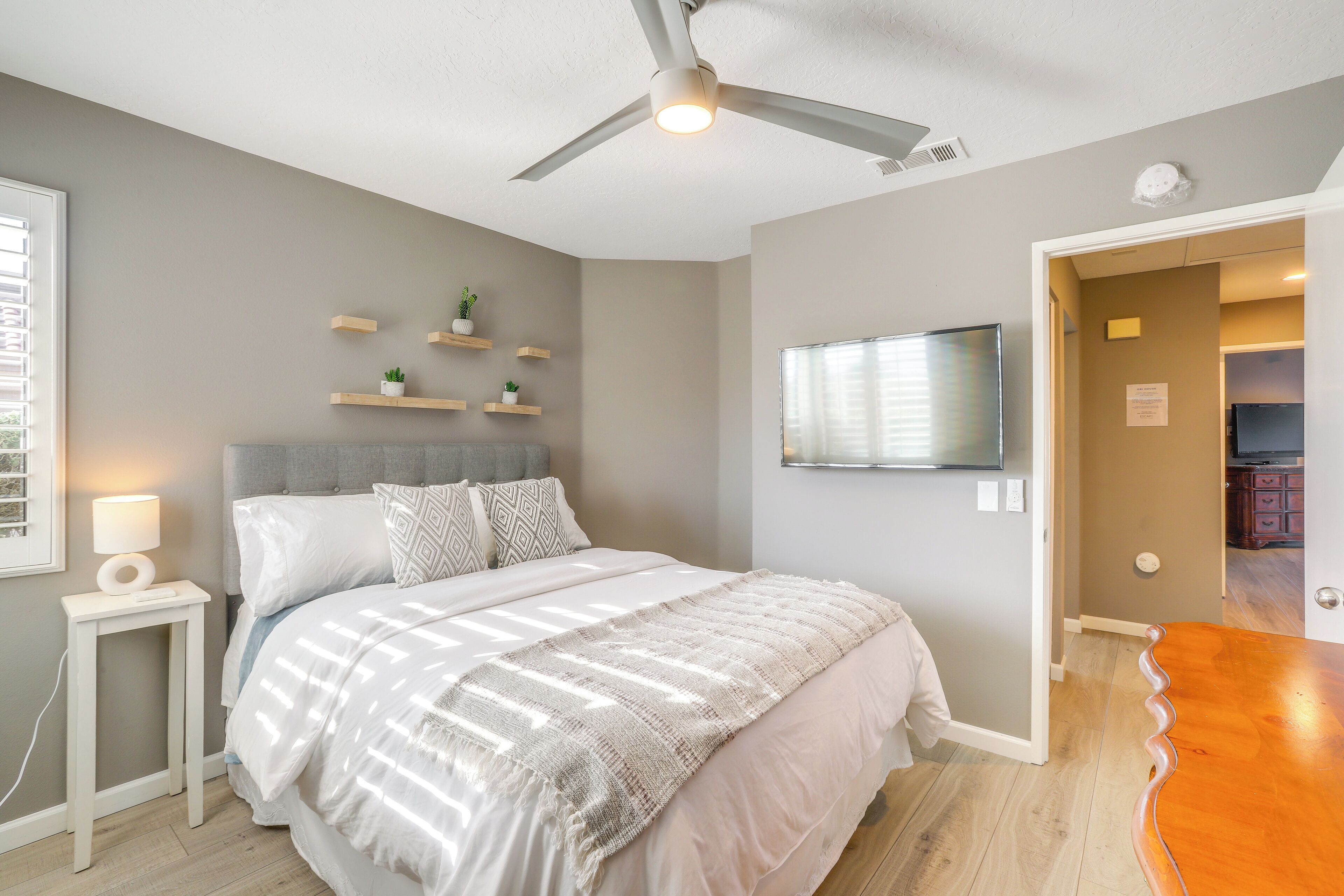 A bedroom with a bed, bedside table, ceiling fan, wall-mounted TV, and built-in shelving with plants.