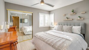 Een slaapkamer met een bed, een plafondventilator, een spiegel, een dressoir en een raam met jaloezieën.