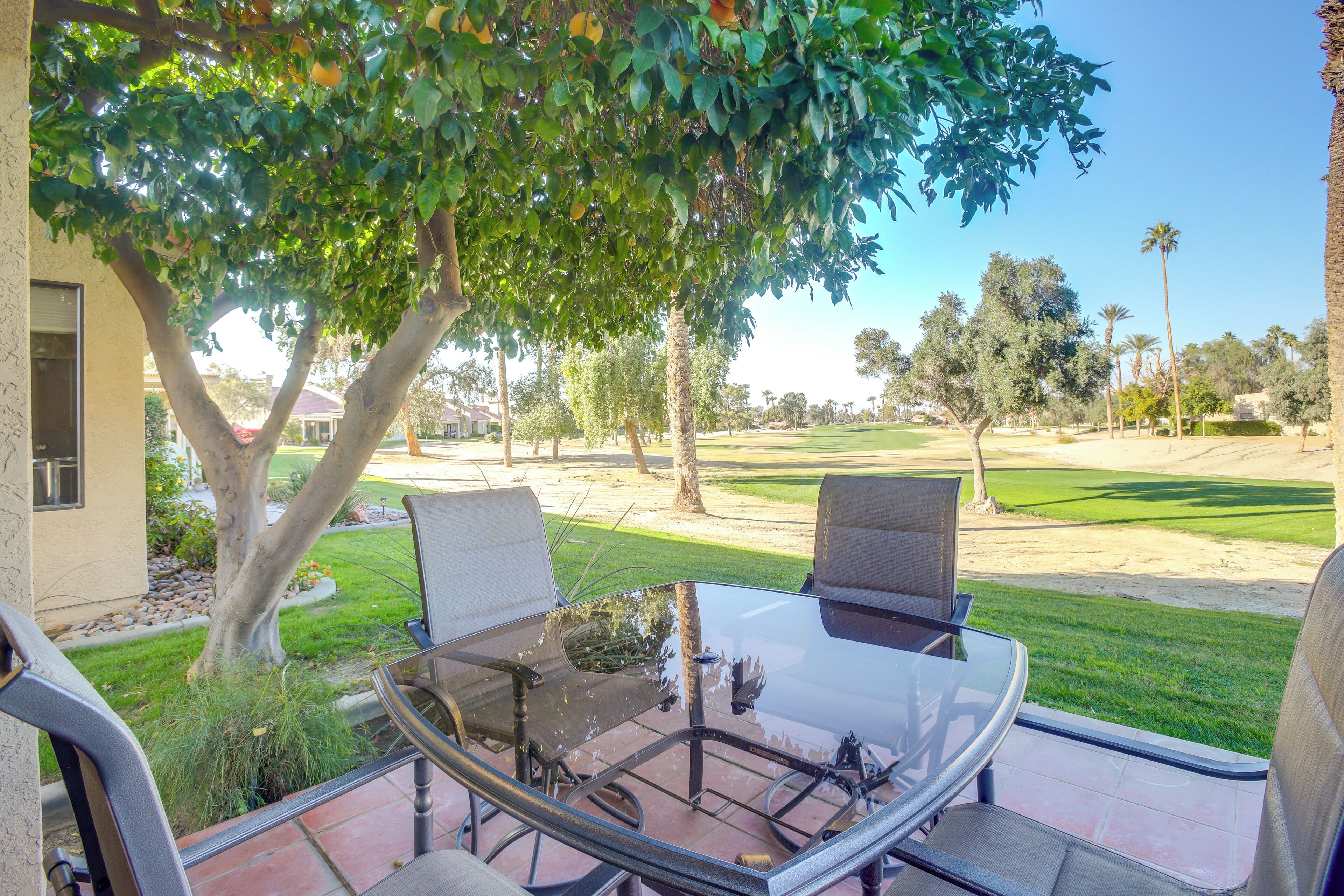 A patio with a glass table and chairs, a view of a golf course, and a tree providing shade.