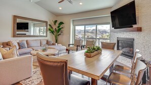 A spacious living room with a fireplace, a large flat-screen TV mounted on a stone wall, and a view of mountains through large windows. - Park City Townhome w/ Hot Tub & Mountain Views! (Kamas)