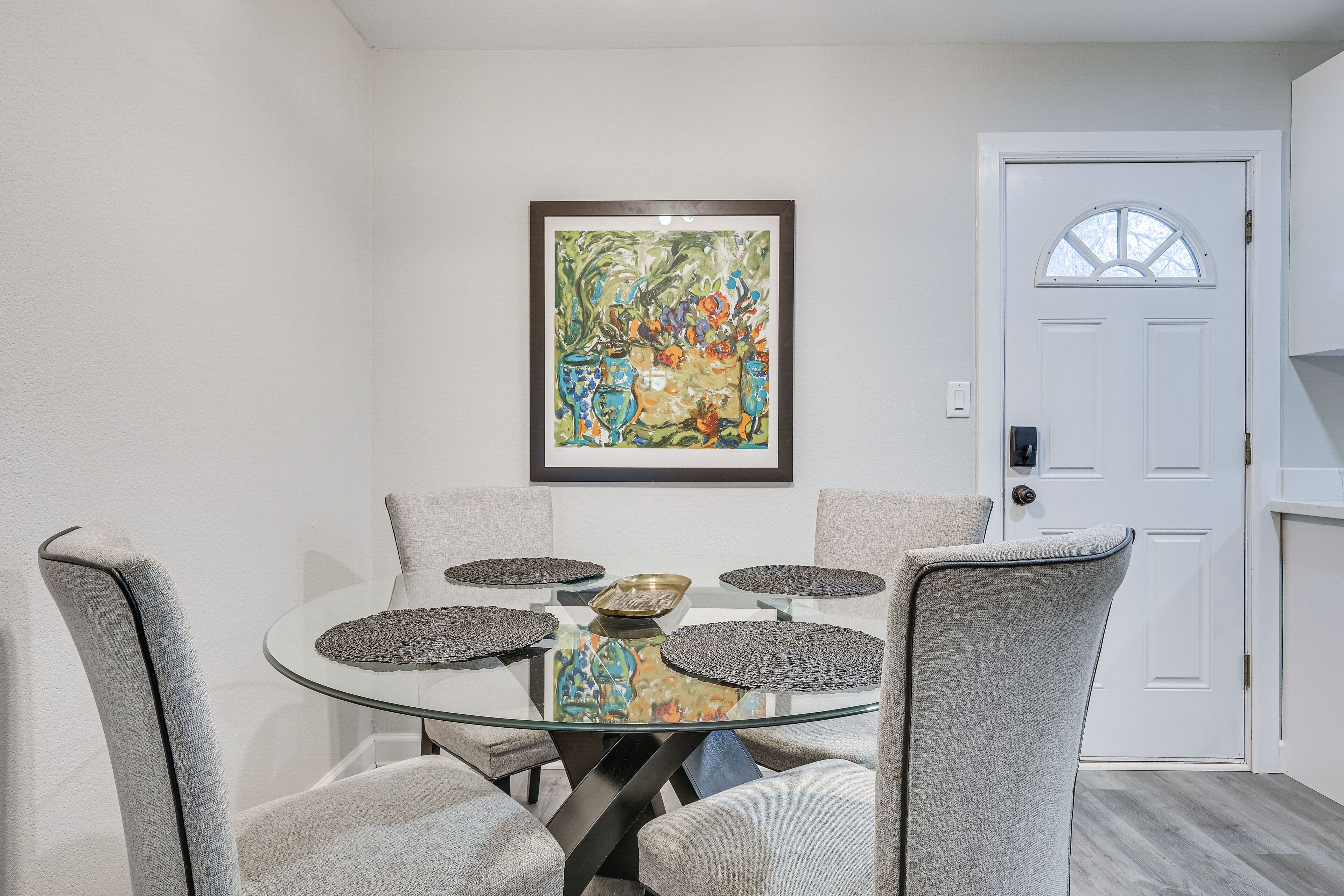 A dining area with a glass table surrounded by grey upholstered chairs, a framed abstract painting on the wall, and a white door with a window.