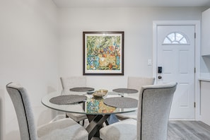 Een eethoek met een glazen tafel, omringd door grijze gestoffeerde stoelen, een wand met een ingelijst abstract schilderij en een witte deur met een raam.