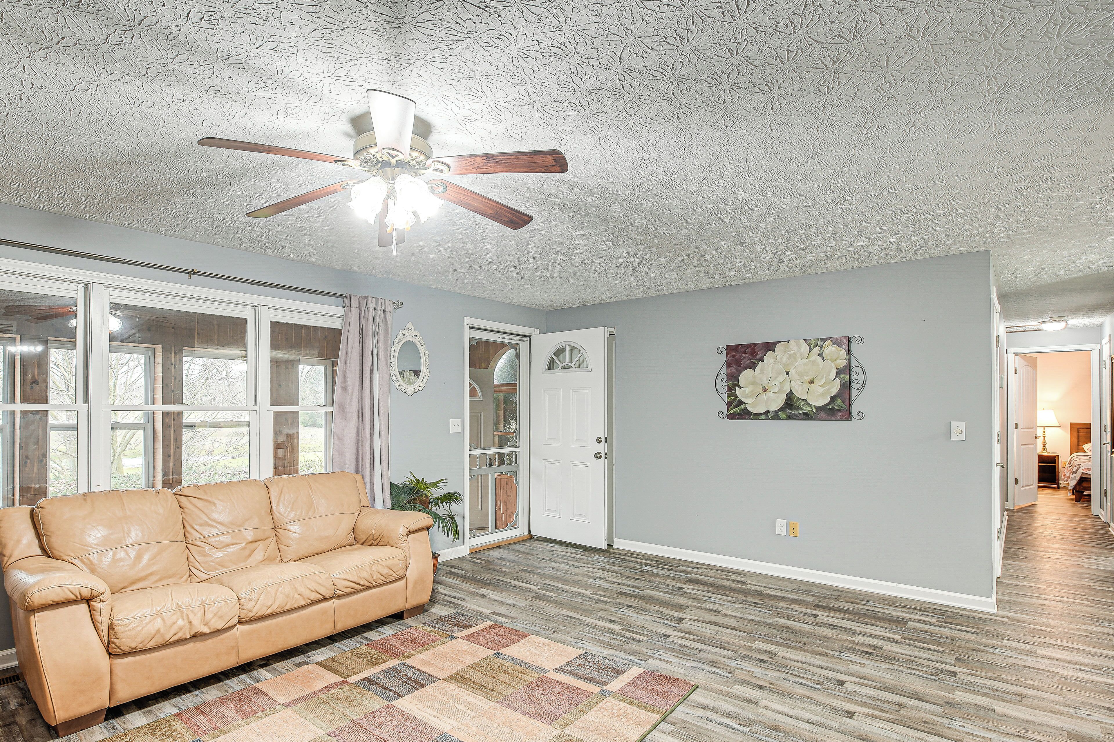 A living room with a beige leather sofa, a patterned rug, a ceiling fan, and a door leading to another room.