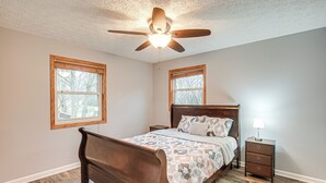Une chambre à coucher avec un lit en bois, deux tables de chevet, un ventilateur de plafond et deux fenêtres.