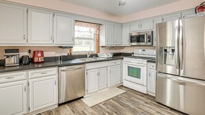 Une cuisine avec des meubles blancs, des appareils en acier inoxydable, un four à micro-ondes, un réfrigérateur et une fenêtre agrémentée d’un rideau à motifs floraux.