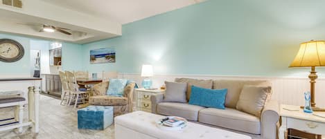 Un salon avec un mur bleu clair, un canapé beige, un pouf bleu, une table à manger avec des chaises, et un coin cuisine à l’arrière-plan.