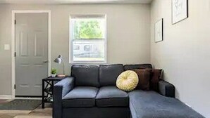 Ein Wohnzimmer mit einem dunkelgrauen Sofa, einem kleinen Beistelltisch mit einer Lampe und einem Fenster mit Blick auf Bäume.