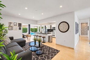 Una sala de estar moderna con sofá, mesa de centro y taburetes de bar. Hay una zona de cocina con salpicadero de ladrillo y vista a una piscina.