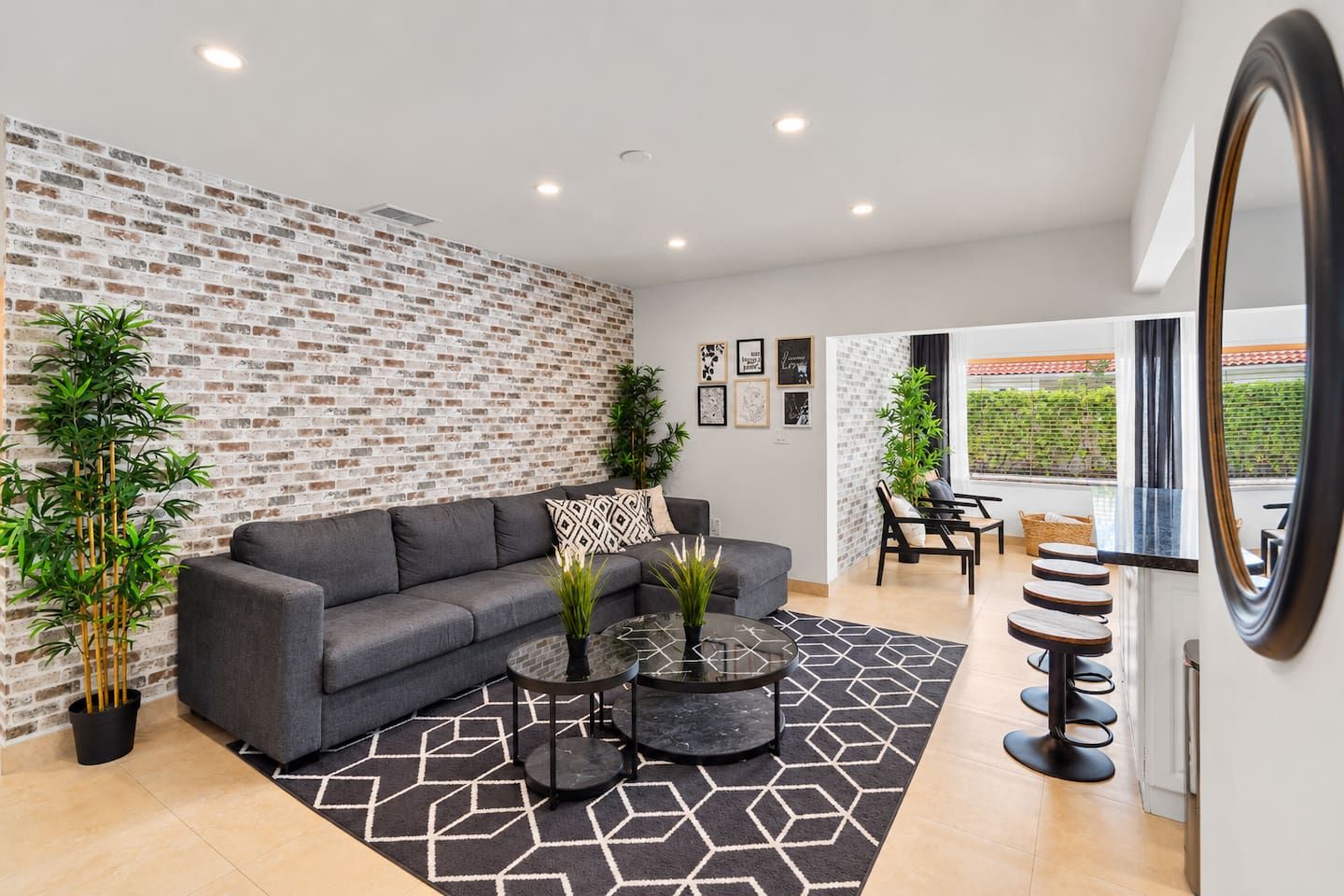 A modern living room with a grey sofa, a coffee table, and a patterned rug. There are two potted plants, a wall with a brick pattern, and a large mirror.