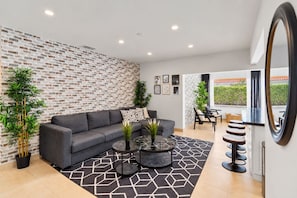 A modern living room with a grey sofa, a coffee table, and a patterned rug. There are two potted plants, a wall with a brick pattern, and a large mirror.