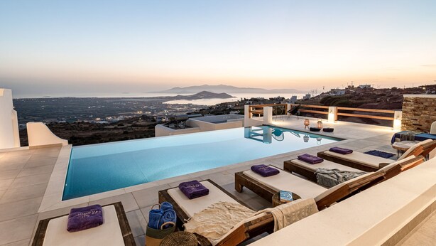 A rooftop infinity pool with lounge chairs and towels, overlooking a coastal cityscape at sunset.