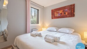 A neatly made bed with white linens and rolled towels, a window with a view of trees, a wall-mounted abstract painting, and a mirror with a wavy design.