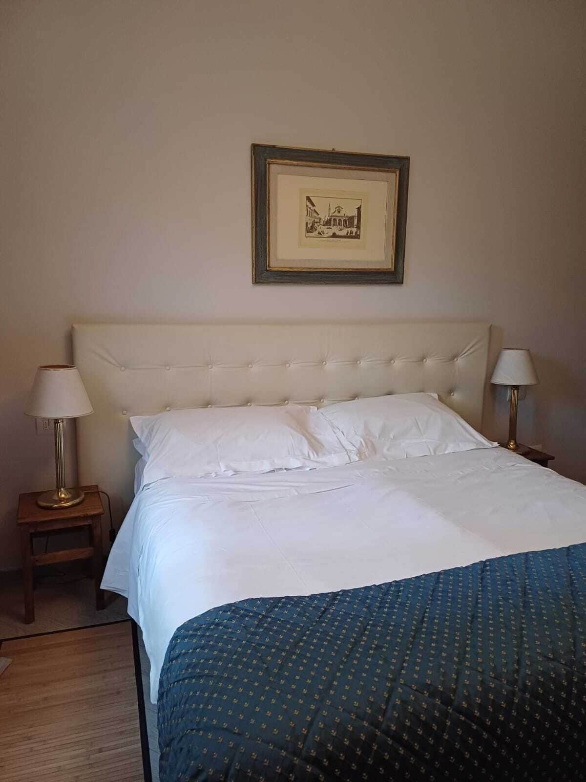 A neatly made bed with white linens, a blue patterned blanket, and two bedside lamps on wooden nightstands. A framed picture hangs above the bed.
