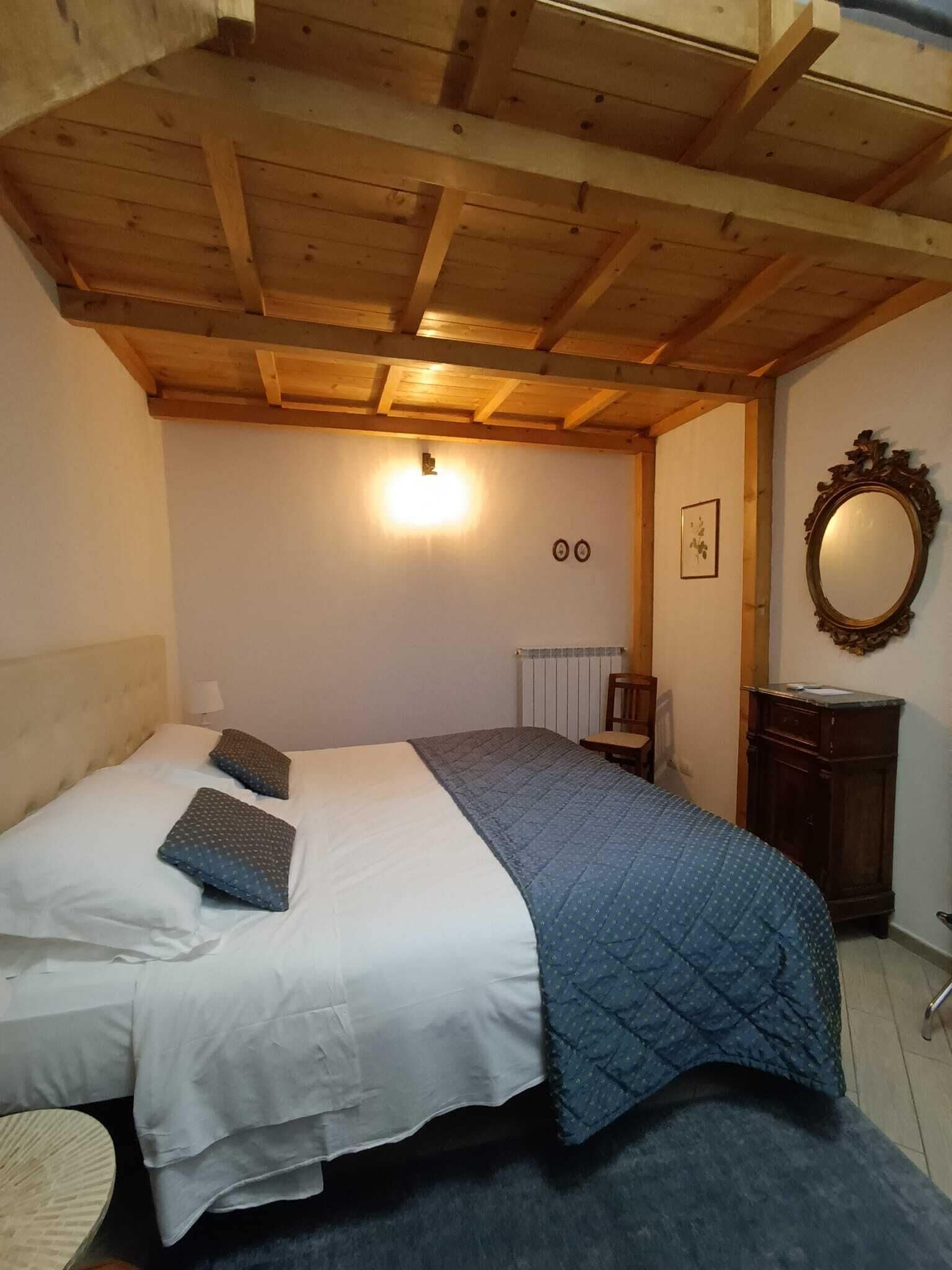 A bedroom with a wooden ceiling, a bed with white and blue bedding, a small table, a chair, a mirror, and a radiator.