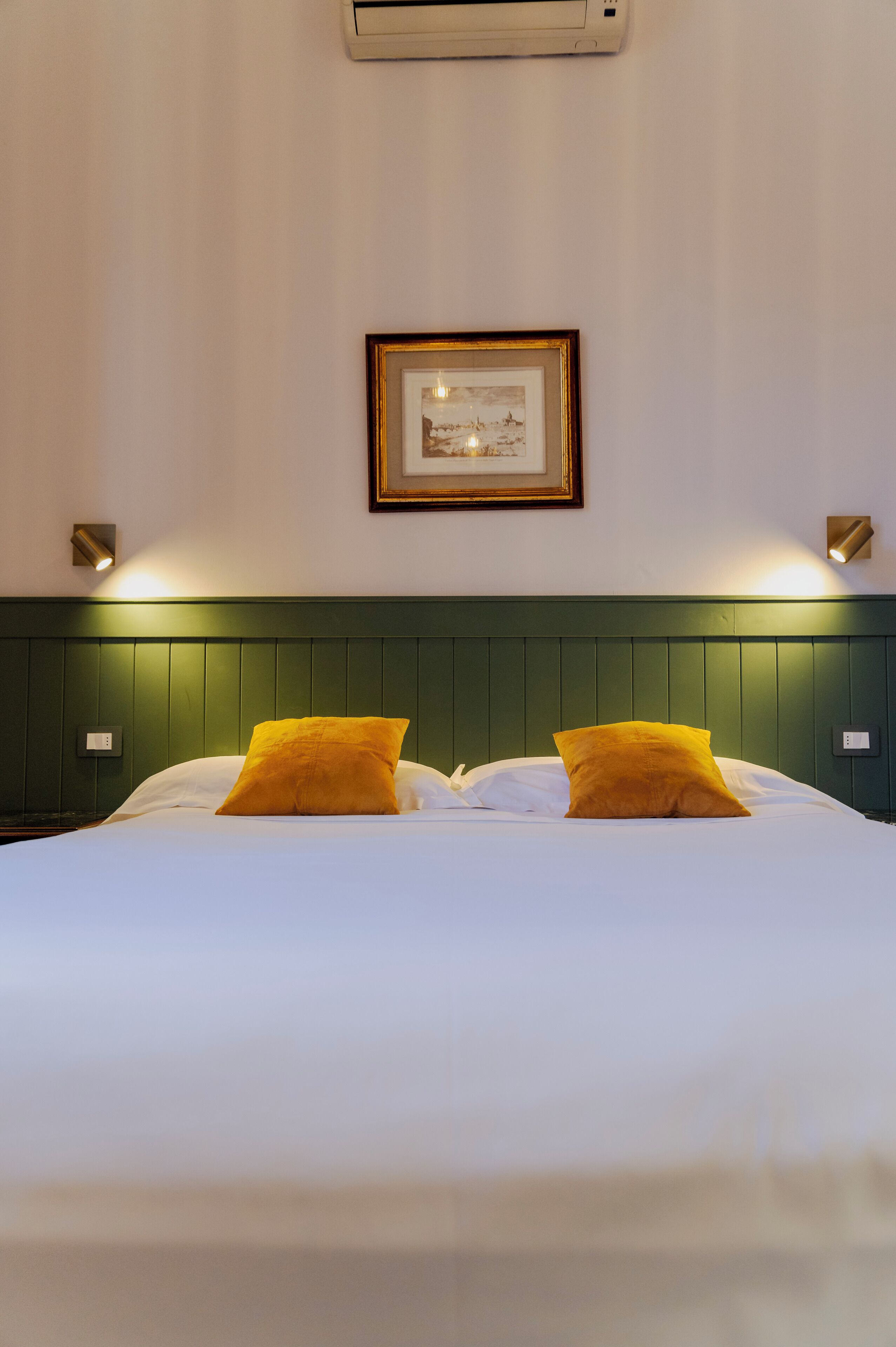 A neatly made bed with white linens and two mustard-colored pillows, a framed picture on the wall, and a green headboard.