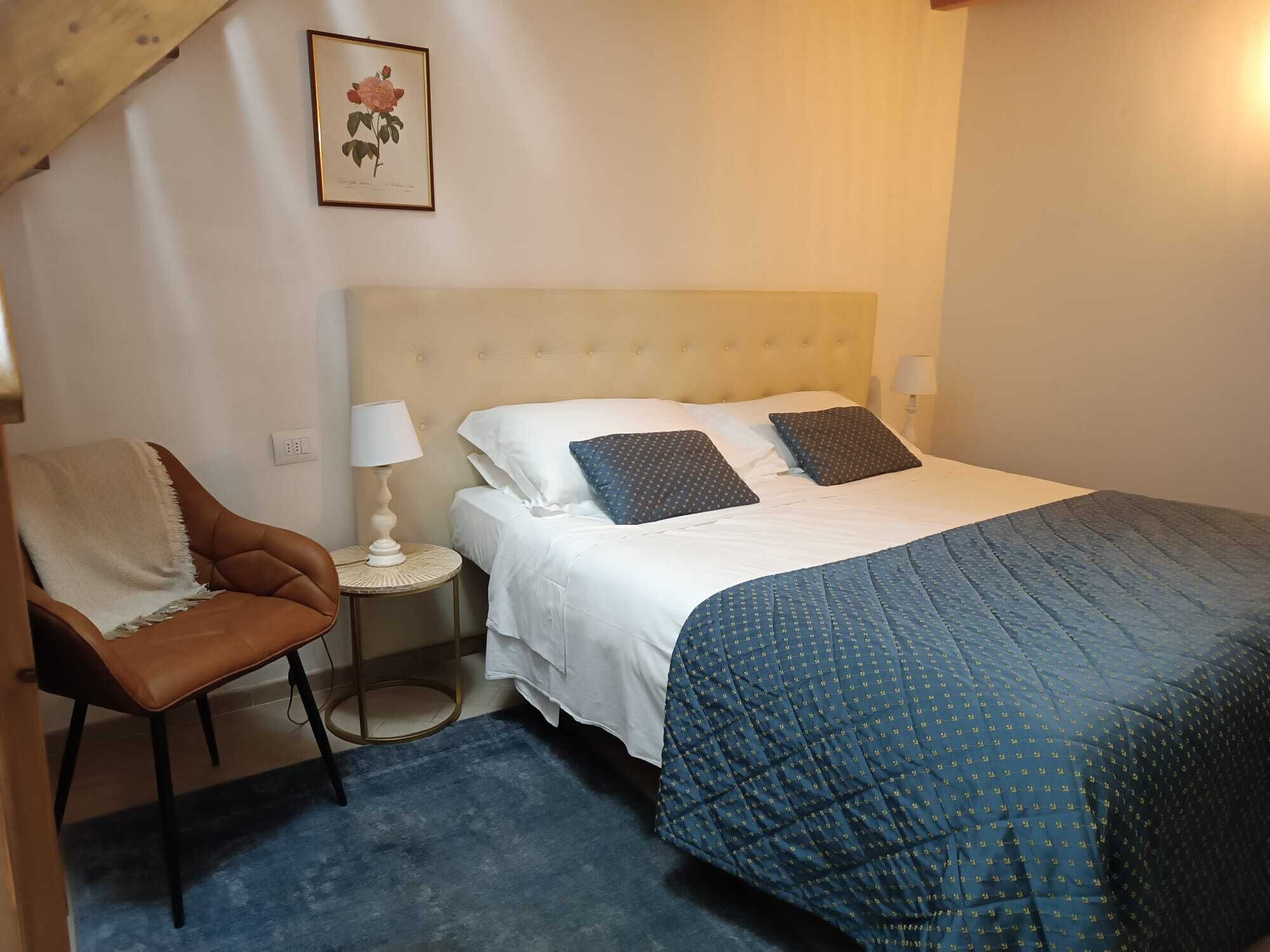 A bedroom with a bed, two pillows, a nightstand with a lamp, a chair, and a framed picture on the wall.