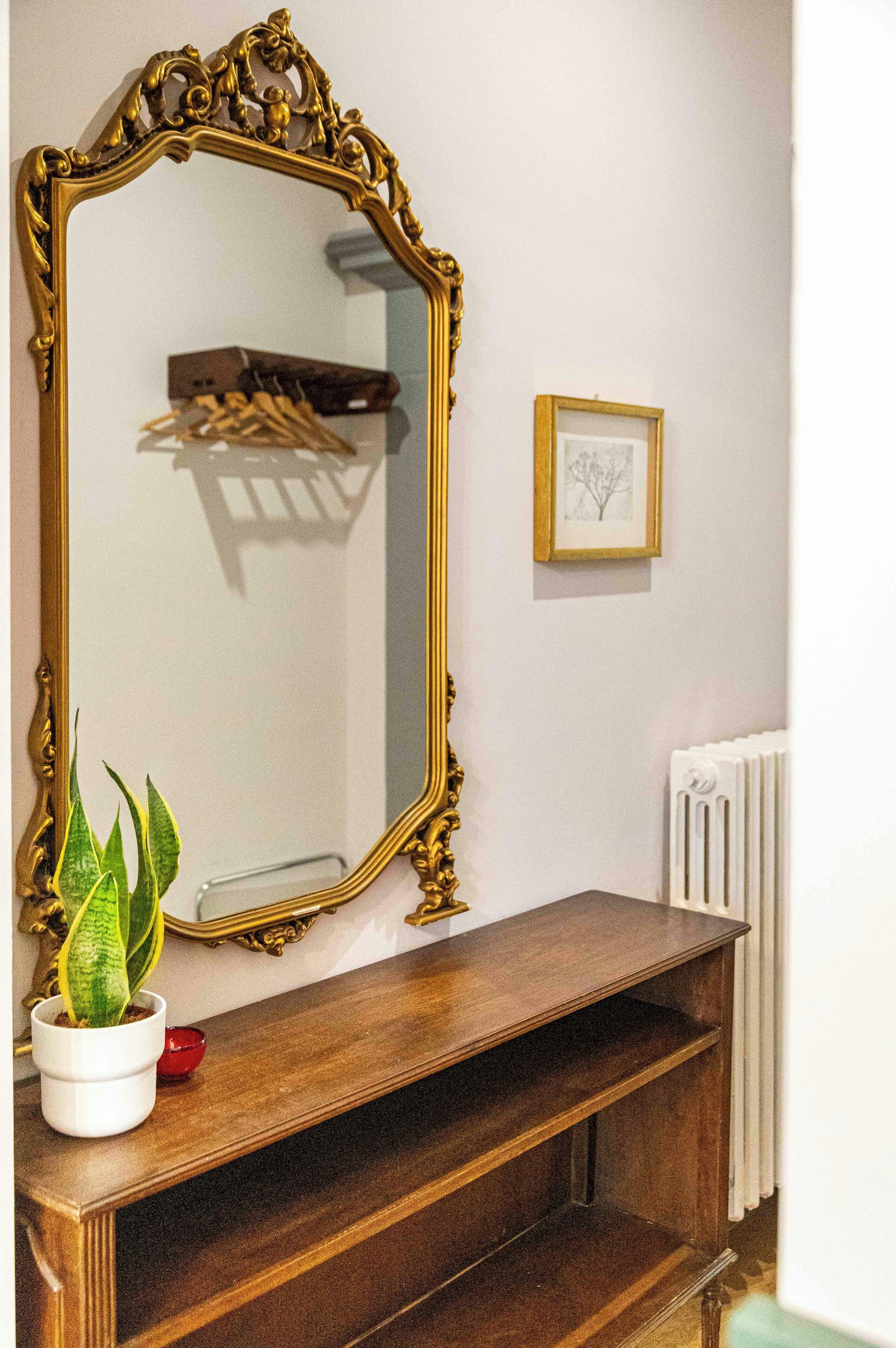Uno specchio con una cornice dorata decorata, un tavolo consolle in legno sottostante, una pianta in vaso e un quadro appeso al muro.