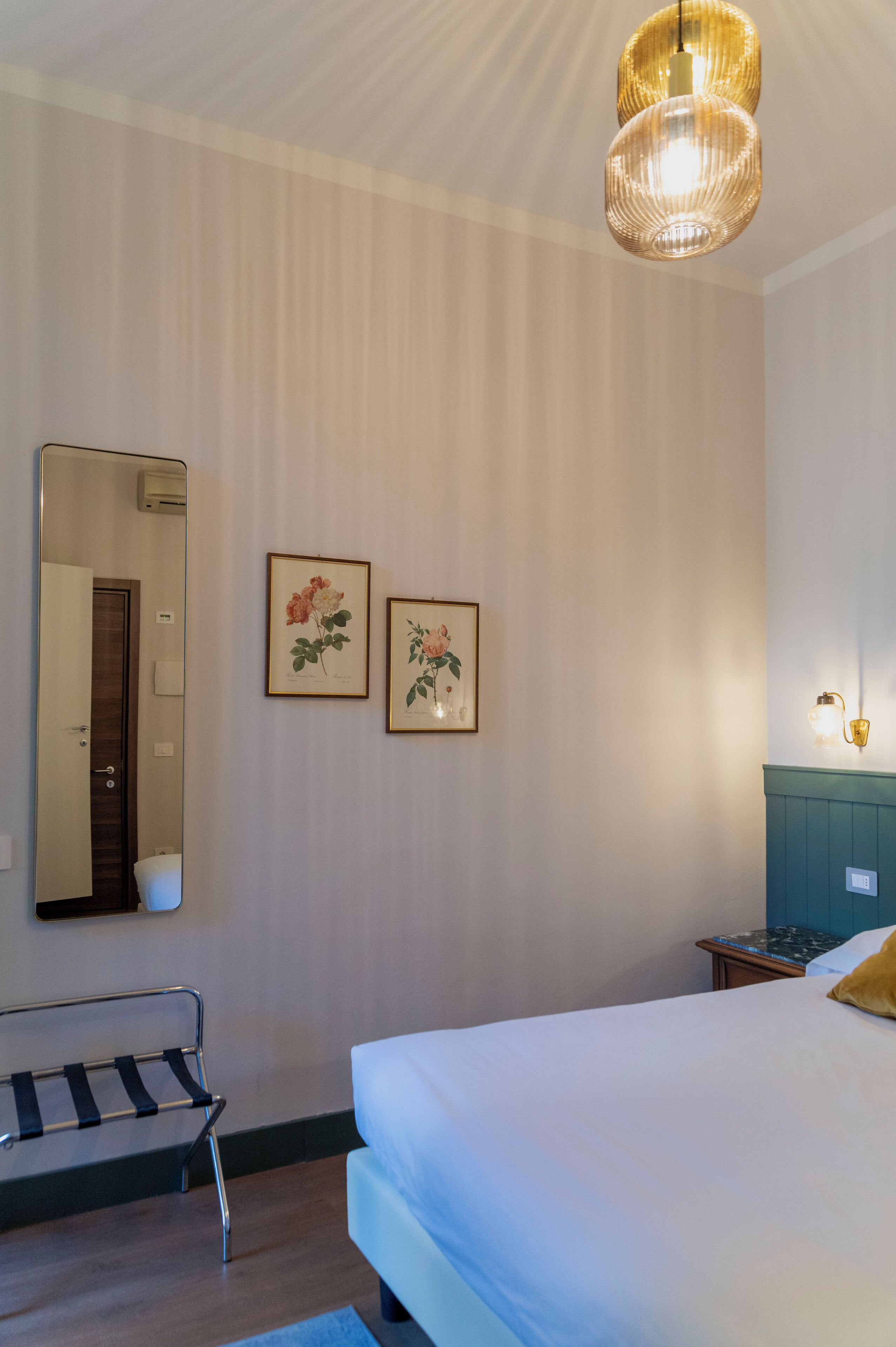 A hotel room with a large bed, a bench, a mirror, two framed floral pictures, and a hanging light fixture.