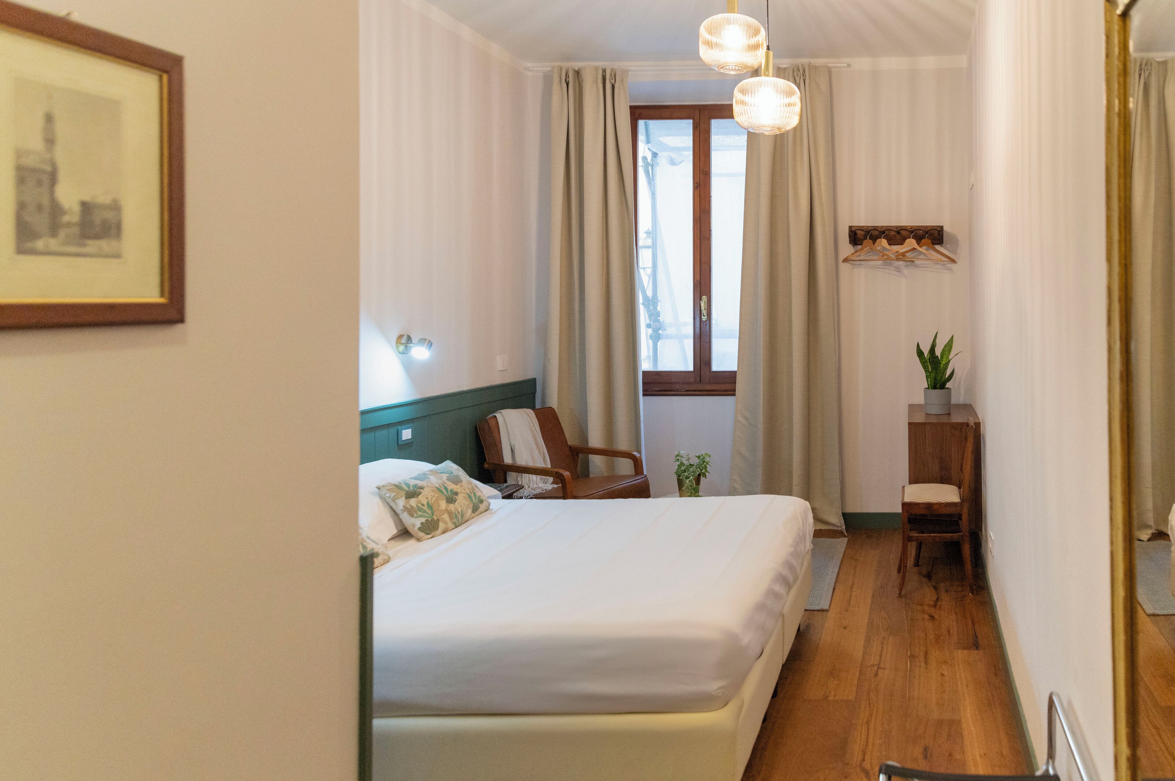 Une chambre d’hôtel avec un lit, une chaise, une petite table avec une plante et une fenêtre avec des rideaux.