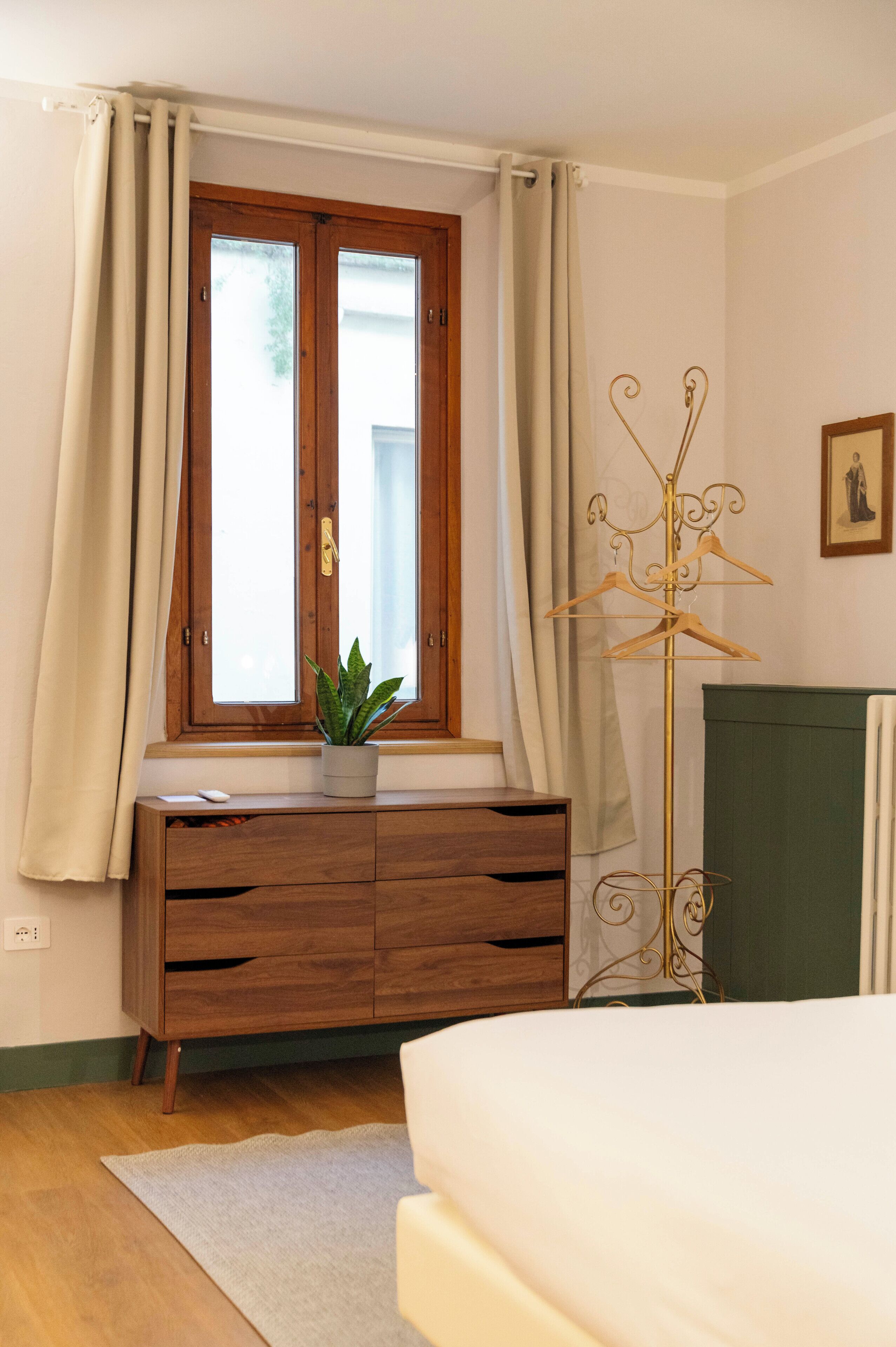 A bedroom with a wooden dresser, a plant on the dresser, a bed, a coat rack, and a window with curtains.