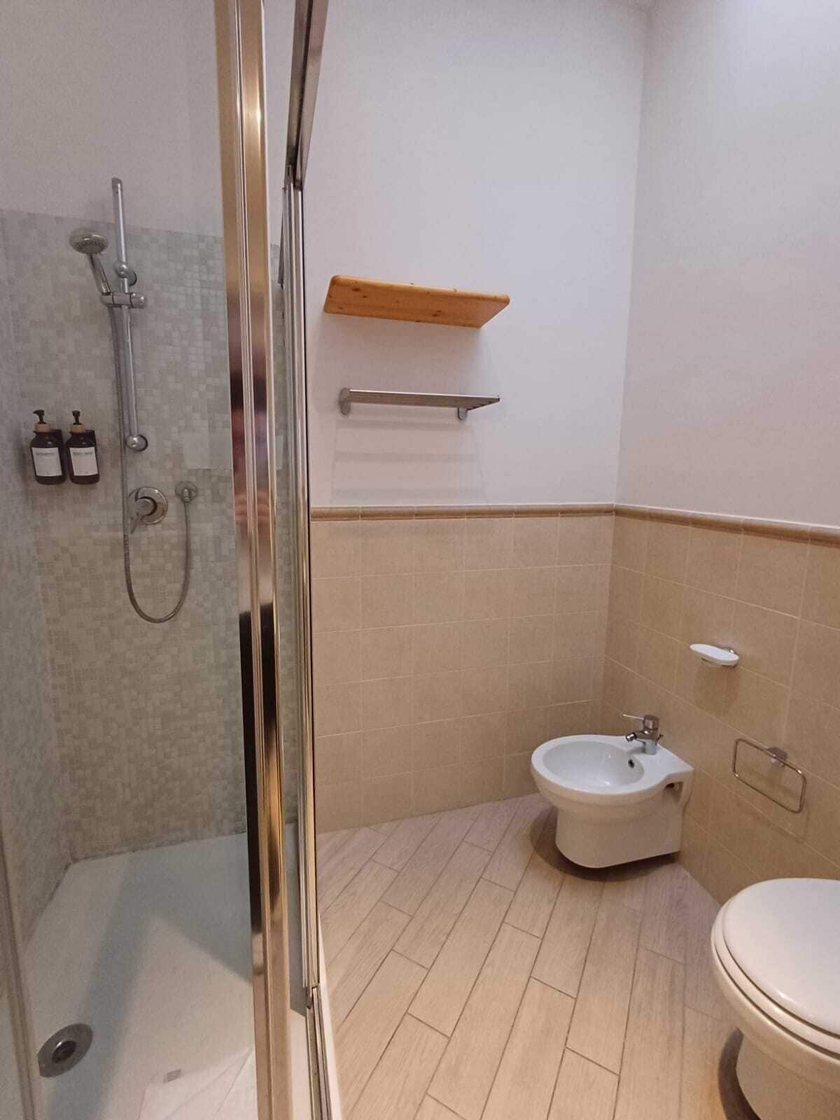 A bathroom with a glass shower enclosure, a bidet, a toilet, and a wooden shelf.
