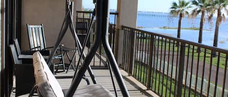 A balcony with a swing chair, a table, and chairs, overlooking a body of water and palm trees.
