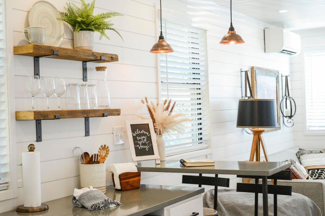 A kitchen with wooden shelves, a hanging pendant light, a wall-mounted air conditioner, and a framed welcome sign.