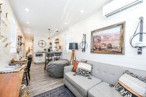 A modern living room with a grey sofa, a dining area with a table and chairs, and a kitchen in the background.