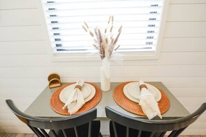 A dining table with two placemats, a vase with dried plants, and a window with blinds. - Luxury Container Home | Pickleball | Rooftop Patio (Waco)