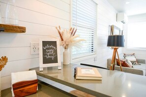 A modern living room with a glass-top coffee table, a framed welcome sign, a vase with pampas grass, a lamp on a tripod, and a sofa with cushions. - Luxury Container Home | Pickleball | Rooftop Patio (Waco)