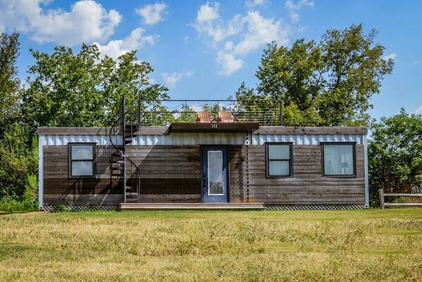 A small, single-story cabin with a wooden exterior, a blue door, and a small porch area. - Luxury Container Home | Pickleball | Rooftop Patio (Waco)