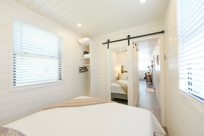 A bedroom with a bed, a window with blinds, a door leading to another room, and a hallway with a table and chairs. - Luxury Container Home | Pickleball | Rooftop Patio (Waco)