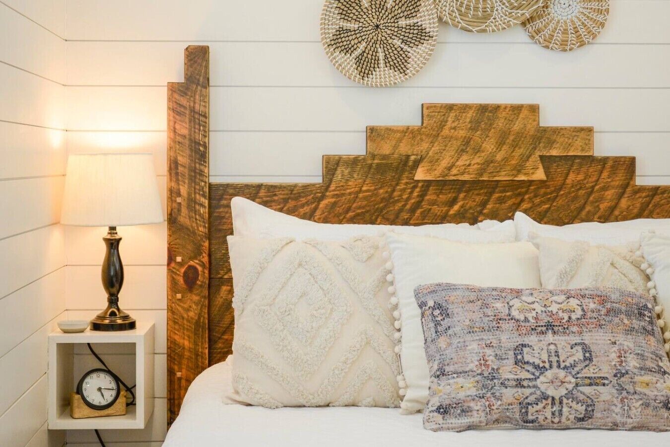 A bed with white and patterned pillows, a wooden headboard, a bedside lamp, and a wall clock.