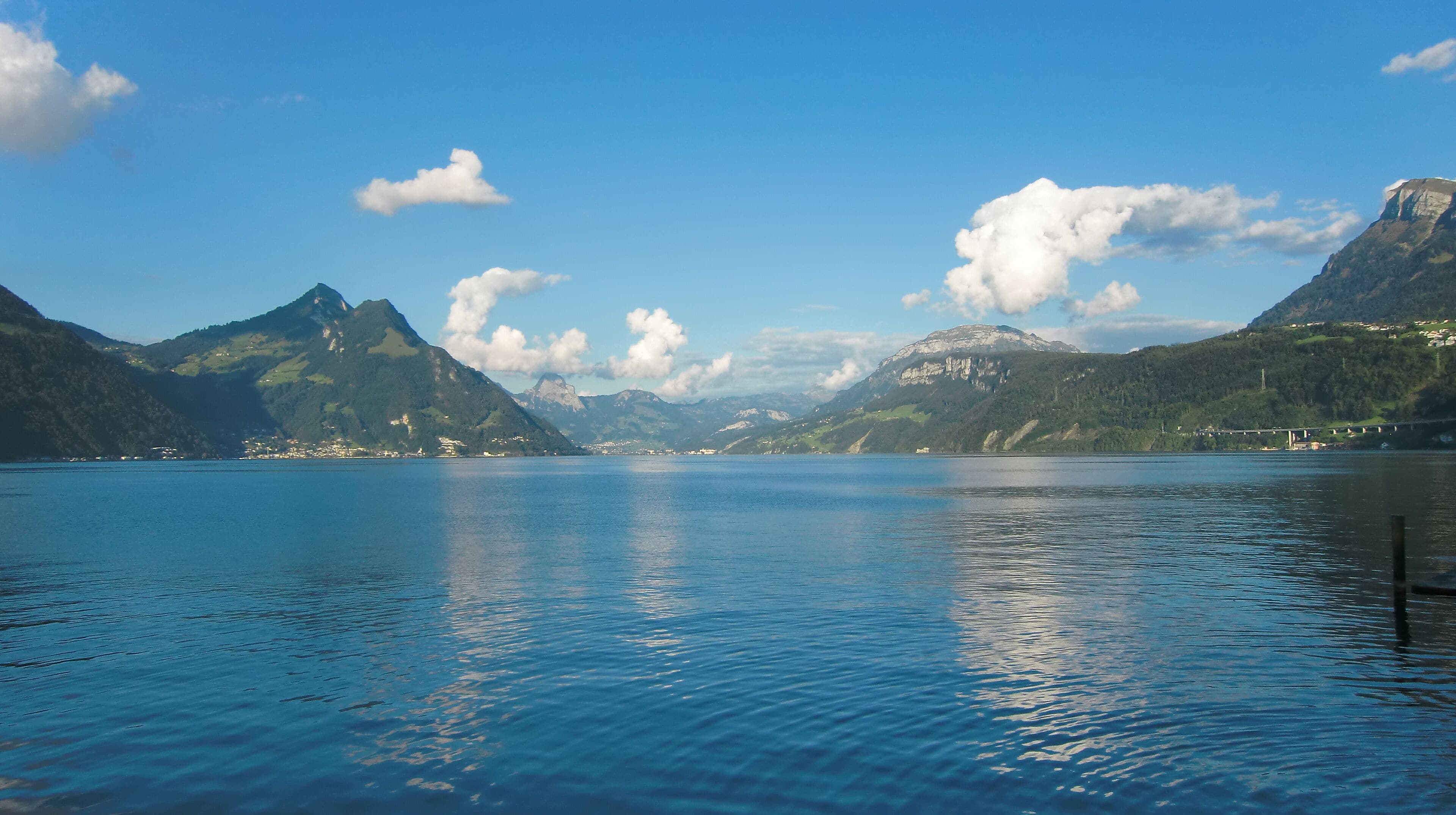 A calm lake surrounded by mountains, with a clear sky and a bridge in the distance.